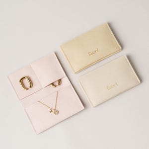 May include: A light pink open jewelry organizer pouch containing a gold chain necklace with a circular pendant and two gold hoop earrings, alongside two closed beige jewelry pouches, each featuring the gold embossed brand name Estsol.