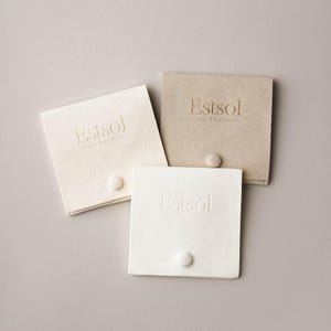 May include: Three square, off-white, beige, and white fabric pouches with the embossed text "Estsol Color Experience". Each pouch has a small, round, white button closure. The pouches are arranged on a neutral gray background.
