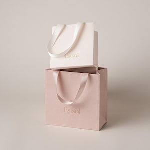 May include: Two stacked gift bags with satin ribbon handles. The top bag is cream-colored with the gold text Estsol, and the bottom bag is a dusty rose color with the gold text Estsol. Both bags feature a smooth, matte finish and matching ribbon.