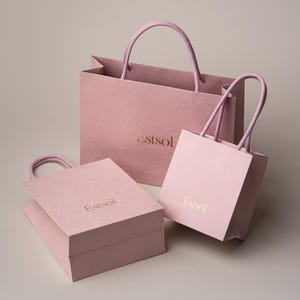 Peut inclure: Trois sacs &agrave; provisions en papier rose de diff&eacute;rentes tailles avec des poign&eacute;es assorties. Les sacs portent le mot "Estsol" imprim&eacute; en or. Les sacs sont sur un fond neutre.
