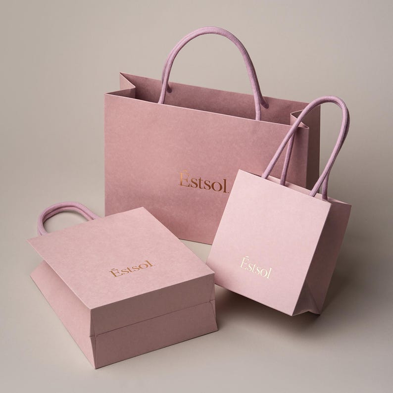Peut inclure: Trois sacs &agrave; provisions en papier rose de diff&eacute;rentes tailles avec des poign&eacute;es assorties. Les sacs portent le mot "Estsol" imprim&eacute; en or. Les sacs sont sur un fond neutre.