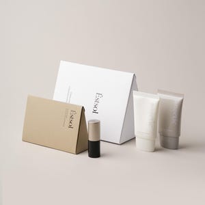 Pu&ograve; includere: Una collezione di prodotti per la cura della pelle che include una grande scatola triangolare bianca, una scatola triangolare color cuoio pi&ugrave; piccola, un contenitore cilindrico con base nera e tappo metallico e due tubi bianchi, tutti con il marchio Estsol.