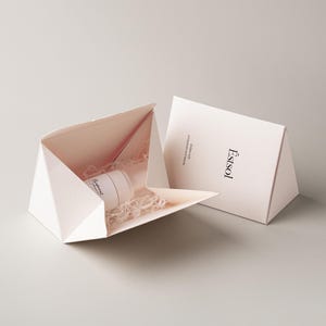 Pu&ograve; includere: Una scatola regalo triangolare rosa pallido con un lembo aperto che rivela un contenitore cilindrico bianco adagiato su trucioli di carta bianca. La scatola presenta il testo Estsol e Endow with uniqueness and beauty stampato in nero.