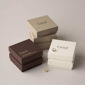 Puede incluir: Una colección de joyeros cuadrados en varios colores: beige, marrón y blanco roto. Cada caja está apilada y presenta el nombre de la marca "Estsol" impreso en la tapa. Dos anillos dorados se colocan cerca de las cajas.