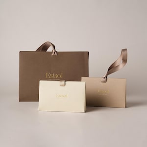 Peut inclure: Un ensemble de trois sacs et boîtes cadeaux dans des tons de brun et de crème. Le plus grand sac est brun foncé, le moyen est brun clair avec une anse en ruban, et le plus petit est une boîte crème avec une languette en ruban, tous avec le texte doré Estsol.