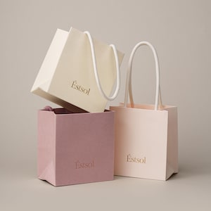 Peut inclure: Trois sacs en papier de diff&eacute;rentes couleurs : cr&egrave;me, mauve et rose p&acirc;le, avec des poign&eacute;es en corde blanche. Les sacs sont empil&eacute;s, le sac cr&egrave;me &eacute;tant au-dessus et le sac mauve au milieu. Le nom de la marque "Estsol" est imprim&eacute; en or sur chaque sac.
