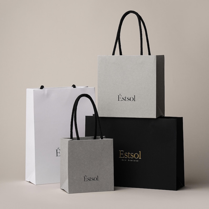 Peut inclure: Quatre sacs de shopping de diff&eacute;rentes tailles et couleurs. Il y a un sac blanc, deux sacs gris et un sac noir. Chaque sac a des poign&eacute;es noires et le texte "Estsol" imprim&eacute; sur le devant. Les sacs sont empil&eacute;s les uns sur les autres.