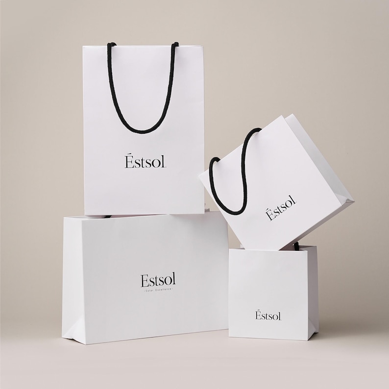 Peut inclure: Quatre sacs en papier blancs de diff&eacute;rentes tailles sont empil&eacute;s. Chaque sac est dot&eacute; d'une poign&eacute;e en corde noire et du mot "Estsol" imprim&eacute; en noir. Les sacs sont dispos&eacute;s sur un fond neutre.