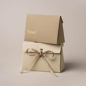 Può includere: Due scatole regalo rettangolari impilate realizzate in cartoncino beige e marrone chiaro. La scatola superiore presenta il testo "Éstsol" in lettere dorate. La scatola inferiore è chiusa con un nastro beige abbinato e mostra il logo "Éstsol".