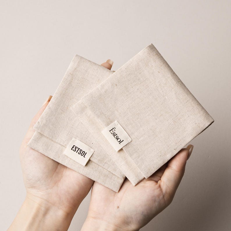 May include: Two beige linen pouches are held in open hands. Each pouch has a small rectangular label with the word "&Egrave;stsol" printed in black. The pouches are folded and appear to be empty.