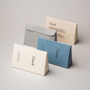 Pu&ograve; includere: Quattro scatole triangolari di carta in color crema, blu e grigio. La scatola color crema presenta il testo 'Estsol' e 'Endow with uniqueness and beauty'. La scatola blu mostra 'Estsol'. La scatola color crema posteriore include 'Estsol' e un paragrafo.