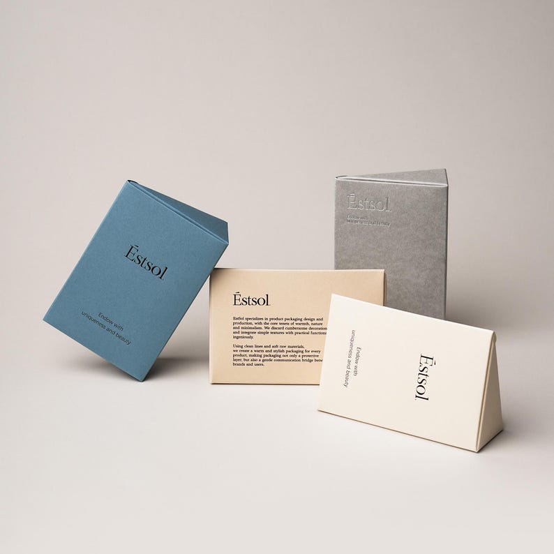 Pu&ograve; includere: Quattro scatole triangolari in cartone di colore blu, beige, crema e grigio. Ogni scatola riporta il nome del marchio Estsol stampato in nero, insieme al testo Endow with uniqueness and beauty. La scatola beige include ulteriore testo descrittivo.