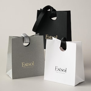 May include: Three square gift bags in black, grey, and white, each featuring a circular cutout handle with a ribbon loop. The black and grey bags display the gold text Estsol, while the white bag features the black text Estsol Solar Excellence.