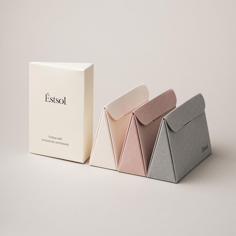 Pu&ograve; includere: Una scatola rettangolare color bianco sporco con il testo Estsol e Endow with uniqueness and beauty, accanto a tre scatole triangolari in crema, rosa antico e grigio, ciascuna dotata di chiusura a patta e nome del marchio Estsol sul lato.