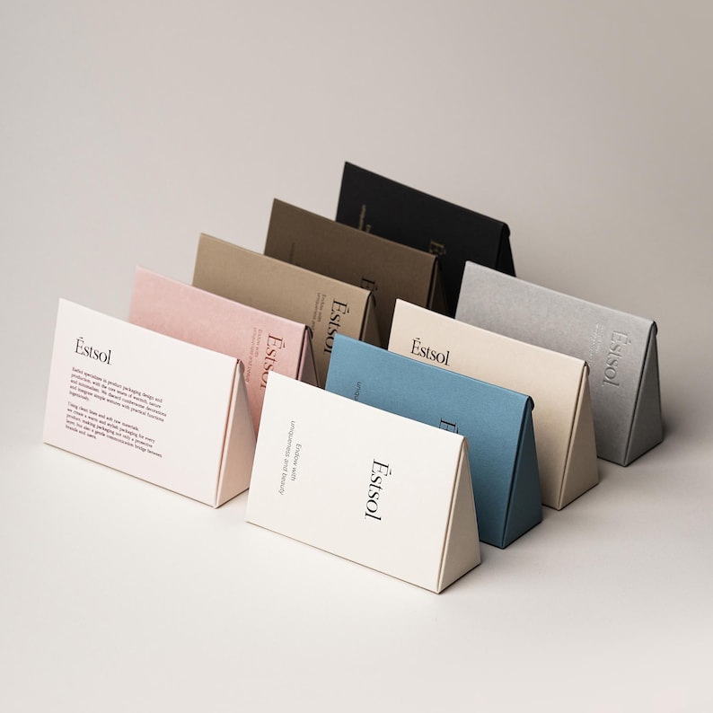 Pu&ograve; includere: Una collezione di scatole di cartone a forma di prisma triangolare in vari colori, tra cui bianco, rosa chiaro, beige, marrone scuro, nero, blu e crema. Ogni scatola presenta il marchio Estsol stampato in nero sulla parte anteriore.