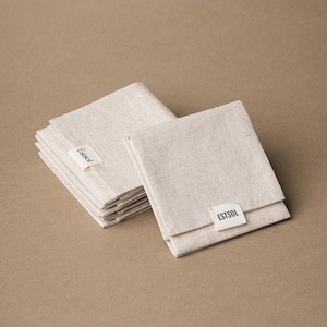 May include: Several folded, beige linen napkins are stacked and arranged on a neutral background. Each napkin has a small white tag with the text "Estsol" or "ESTSOL" printed in black. The napkins appear to be new and unused.