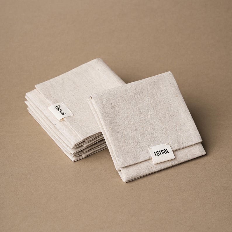 May include: Several folded, beige linen napkins are stacked and arranged on a neutral background. Each napkin has a small white tag with the text "Estsol" or "ESTSOL" printed in black. The napkins appear to be new and unused.