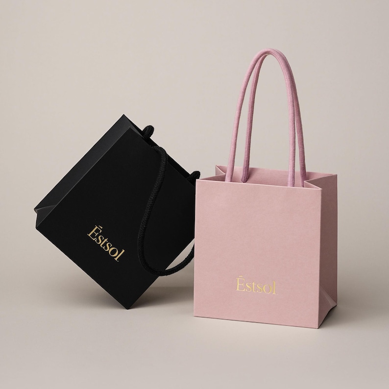 Peut inclure: Deux sacs cadeaux &eacute;l&eacute;gants. L'un est noir avec une poign&eacute;e en corde noire, l'autre est rose avec des poign&eacute;es en corde rose. Les deux sacs portent le texte dor&eacute; "Estsol" imprim&eacute; sur le devant.