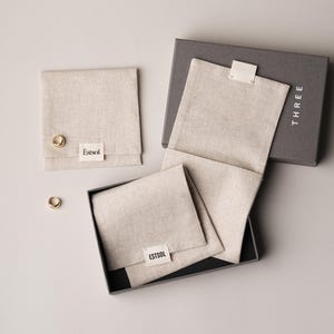 May include: A set of linen jewelry pouches and a gray box. The pouches are beige with the brand name "&Eacute;stsol" on a small white label. A pair of gold earrings is visible. The box is labeled "THREE".