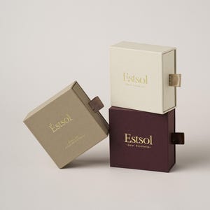Peut inclure: Trois boîtes carrées empilées, beige, crème et bordeaux. Chaque boîte a une tirette et un texte doré qui dit "Estsol". La boîte beige est inclinée, révélant le texte "Estsol - Solar Excellence".