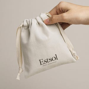 May include: A beige drawstring pouch with the word "Estsol" printed in dark brown. The pouch is held by a hand and has a matching beige drawstring closure. The text "-Solar Excellence-" is printed below the brand name.