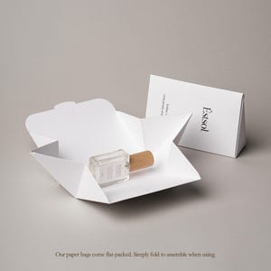 Pu&ograve; includere: Un flacone rettangolare in vetro con tappo in legno chiaro riposa all'interno di una scatola di carta bianca aperta. Accanto, una confezione di carta bianca piegata mostra il testo Estsol e Endow uniqueness and. Gli oggetti sono su sfondo neutro.