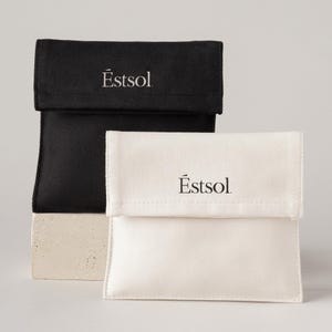 May include: Two rectangular fabric pouches with fold-over flaps, one in solid black and one in off-white. Each pouch features the brand name Éstsol printed in a centred, serif font on the front flap. The pouches have a smooth, matte texture.