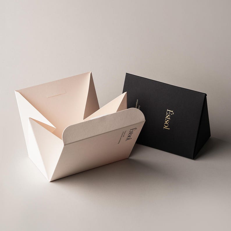 Pu&ograve; includere: Una scatola di carta color crema chiaro con un design pieghevole geometrico e una scatola di carta nera abbinata posizionata in verticale accanto. Entrambe le scatole presentano il marchio Estsol stampato in un carattere serif dorato.