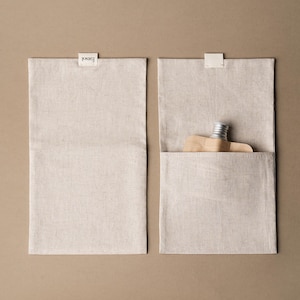 May include: Two rectangular, beige linen pouches with a small, white label at the top. One pouch is closed, the other open, revealing a small, silver-capped bottle and a wooden object. The label reads "Essol".