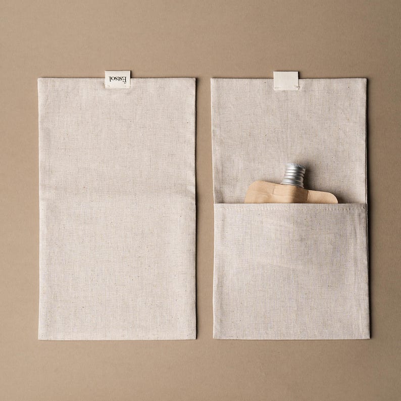 May include: Two rectangular, beige linen pouches with a small, white label at the top. One pouch is closed, the other open, revealing a small, silver-capped bottle and a wooden object. The label reads "Essol".