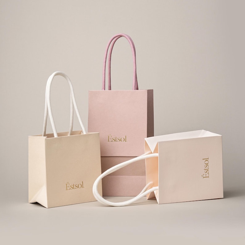 Peut inclure: Trois petits sacs cadeaux carr&eacute;s en papier beige et rose, avec des poign&eacute;es assorties. Les sacs portent le mot "Estsol" imprim&eacute; en or. Un sac est couch&eacute; sur le c&ocirc;t&eacute;.