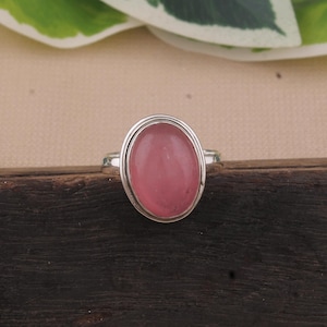 May include: A silver ring featuring a large, oval-shaped pink gemstone. The gemstone is set within a silver bezel, and the ring band is also silver. The ring is displayed on a dark wooden surface.