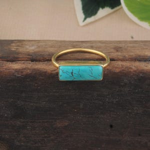 May include: A gold-toned ring featuring a rectangular, horizontally oriented turquoise stone. The stone is bright blue with dark, thin, irregular veining throughout its surface. The metal band is thin, smooth, and circular, framing the stone setting.
