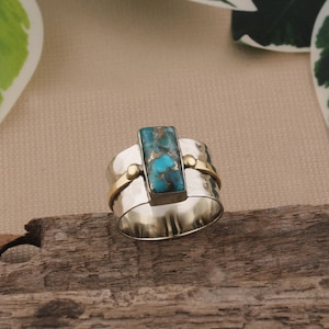 May include: A wide silver ring with a rectangular turquoise stone at the center. The ring has gold accents and a hammered texture. The turquoise stone has a mix of blue and brown colors.