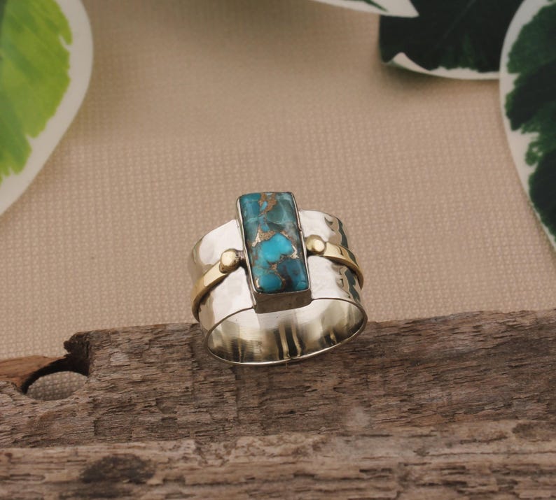 May include: A wide silver ring with a rectangular turquoise stone at the center. The ring has gold accents and a hammered texture. The turquoise stone has a mix of blue and brown colors.