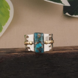 May include: A silver ring with a rectangular turquoise stone at the center. The ring has a hammered texture and is accented with gold-colored bands and small spheres. The ring is displayed on a dark wooden surface.