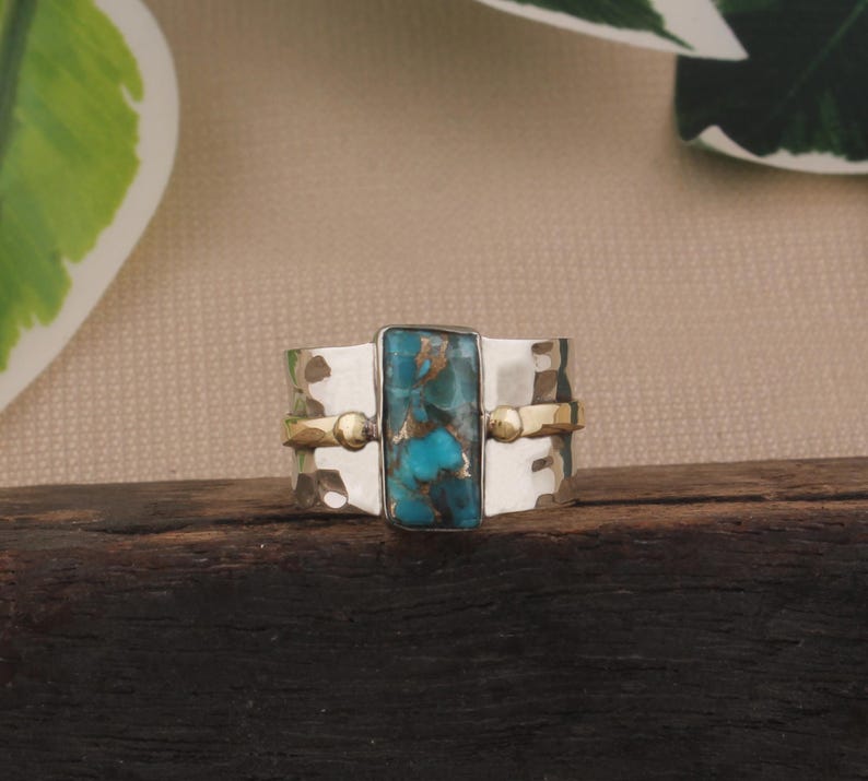 May include: A silver ring with a rectangular turquoise stone at the center. The ring has a hammered texture and is accented with gold-colored bands and small spheres. The ring is displayed on a dark wooden surface.