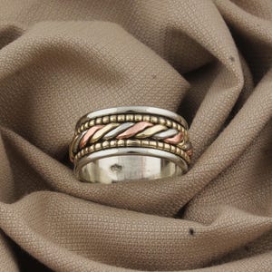 May include: A silver ring with a multi-tone design. The ring features a central band of woven gold, rose gold, and silver, with a beaded border. The ring is set against a neutral fabric background.