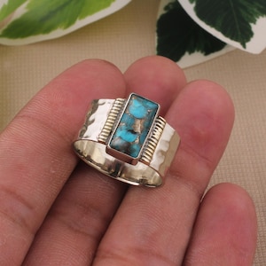 May include: A silver ring with a rectangular turquoise stone. The ring has a hammered texture on the band and gold-colored accents. The turquoise stone has a mix of blue and brown hues.