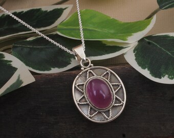 May include: A silver pendant necklace featuring an oval, deep pink gemstone. The gemstone is set within a starburst design, surrounded by a twisted rope border. The pendant hangs from a delicate silver chain.