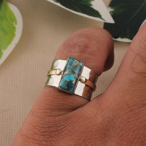 May include: A silver ring with a rectangular turquoise stone. The ring has gold accents and a hammered texture. The turquoise stone is a vibrant blue-green color, and the ring is worn on a finger.