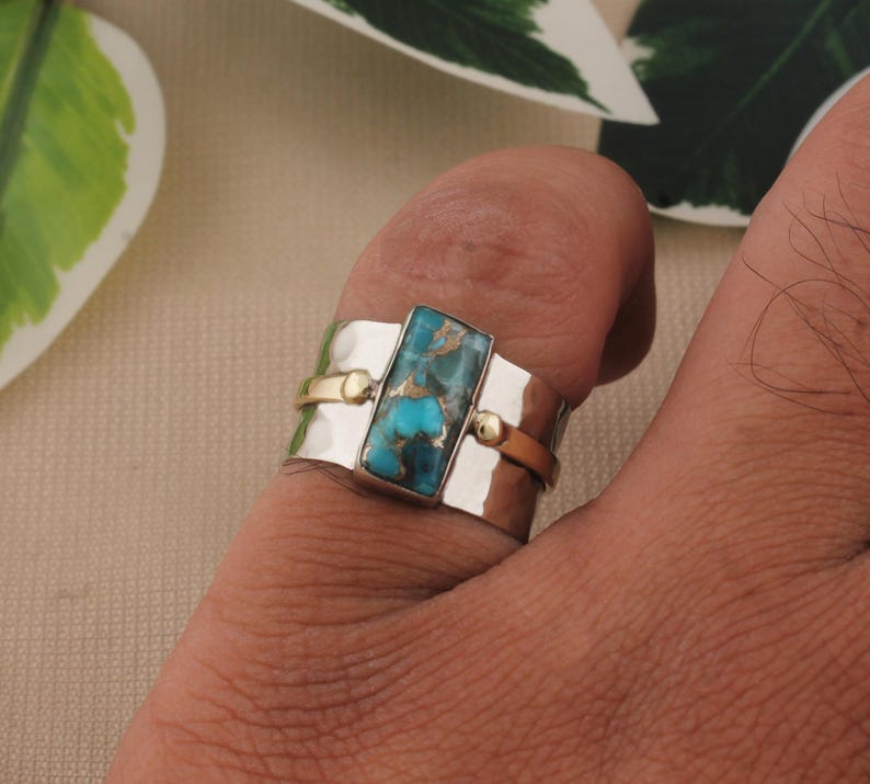 May include: A silver ring with a rectangular turquoise stone. The ring has gold accents and a hammered texture. The turquoise stone is a vibrant blue-green color, and the ring is worn on a finger.
