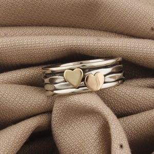 Puede incluir: Un anillo de plata con dos detalles de corazón dorado. El anillo tiene un diseño de banda texturizada y está sobre un fondo de tela neutra. Los corazones son de diferentes tonos dorados.