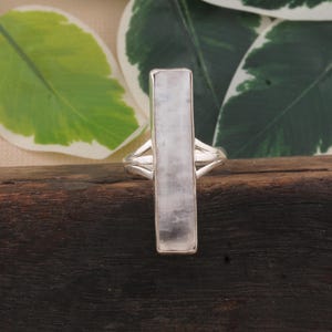 Peut inclure: Une bague en argent avec une pierre rectangulaire de couleur claire. L'anneau de la bague a un design divisé de chaque côté de la pierre. La pierre est de couleur gris clair avec de subtiles variations. La bague est posée sur une surface en bois sombre.