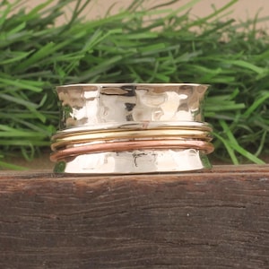 May include: A silver spinner ring with a hammered texture. The ring has three bands in gold, copper, and silver. The ring is displayed on a wooden surface with green foliage in the background.