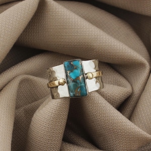 May include: A silver ring with a rectangular turquoise stone at the center. The ring has a hammered texture and is accented with gold-colored bands and small spheres. The turquoise stone has a mix of blue and brown hues.