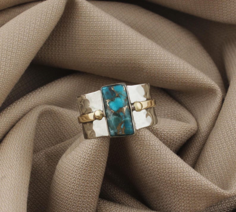 May include: A silver ring with a rectangular turquoise stone at the center. The ring has a hammered texture and is accented with gold-colored bands and small spheres. The turquoise stone has a mix of blue and brown hues.