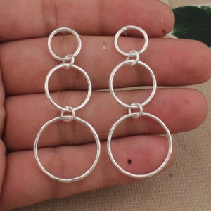 Könnte beinhalten: Ein Paar silberne Ohrringe mit einem Design aus verbundenen Kreisen. Jeder Ohrring hat drei unterschiedlich große Kreise, die durch kleine Schlaufen verbunden sind. Die Ohrringe werden vor einem neutralen Hintergrund gezeigt.