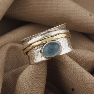 May include: A silver ring with a gold band and a light blue oval gemstone. The ring has a textured design and is set against a brown fabric background.