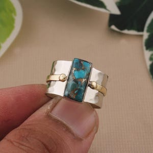May include: Silver ring with a rectangular turquoise stone. The ring has gold accents and a hammered texture on the silver band. The turquoise stone has a mix of blue and brown colors.